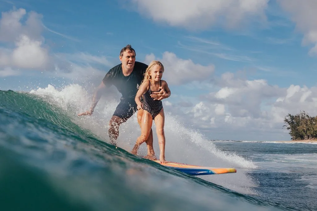 Chris riding a wave with his daughter standing on the board in front of him